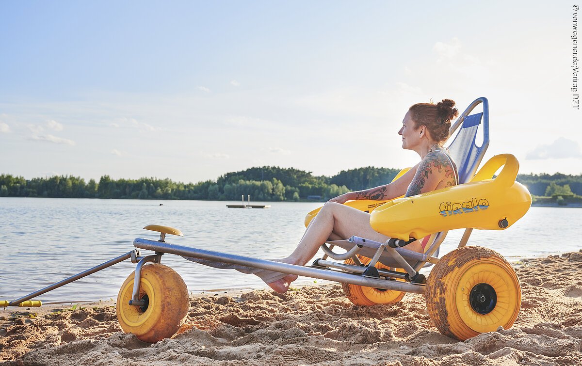 Strandrollstuhl am Großen Brombachsee Blick auf Sandstrand und Wasser. Eine Frau sitzt im geländegeängigen Strandrollstuhl und blickt auf den Großen Brombachsee.
