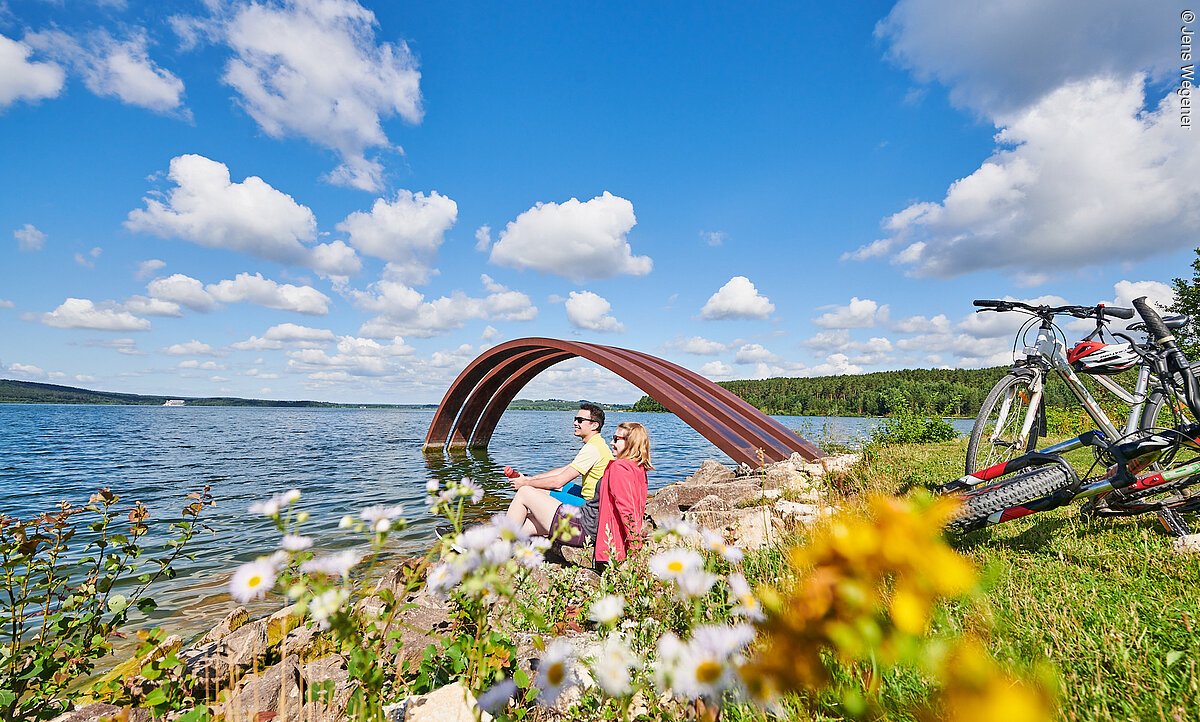 Radeltour am See Ein junges Pärchen macht eine Pause vom Radeln am Ufer eines Sees. Sie sitzen direkt am Wasser auf einer grünen Wiese. Hinter ihnen stehen Fahrräder. Im Wasser steht ein roter Metallbogen.
