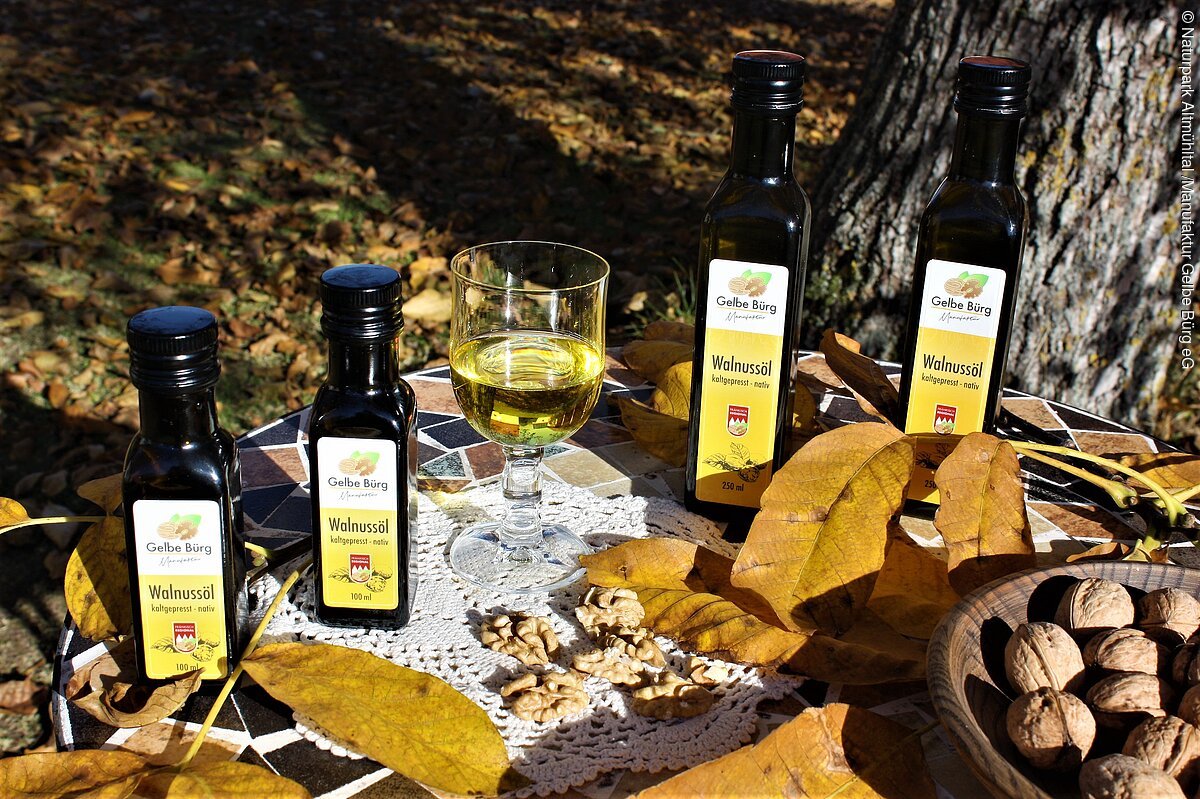 Walnussölauswahl Manufaktur Gelbe Bürg Flaschen mit Walnussöl, ein Glas Öl, Walnüsse und Herbstblätter auf einem Tisch im Freien.