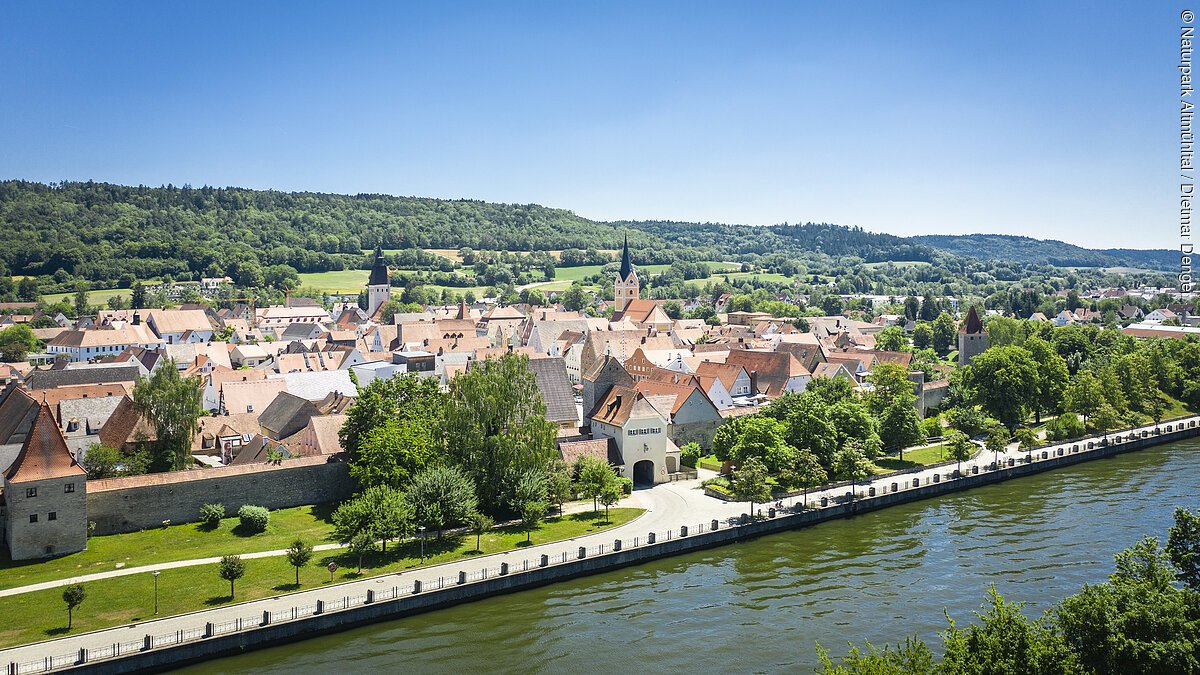 Berching Blick auf eine Stadt mit roten Dächern, Flussufer, Bäumen und bewaldeten Hügeln im Hintergrund bei klarem Himmel.