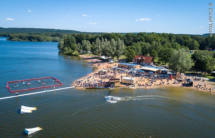 Wakeboardanlage Luftbild von der Wakeboardanlage. Im See Hindernisse für die Wakeboardfahrer. Am See ein Strand mit vielen Besuchern. Dahinter grüne Bäume und Sträucher.