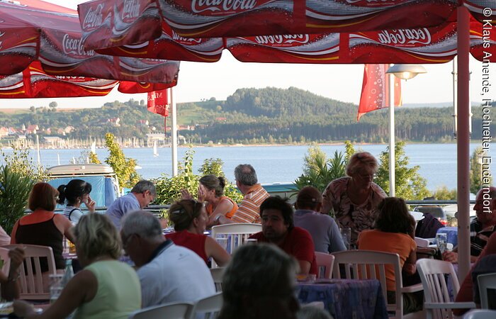 Blick vom barrierefreien Außenbereich des Cafe-Restaurants „Zum Hochreiter“ auf den Großen Brombachsee im Spalter Ortsteil Enderndorf Gäste sitzen in einem Biergarten mit weißen Stühlen und roten Coca-Cola-Schirmen. Im Hintergund wird der Brombachsee von der Sonne beleuchtet.