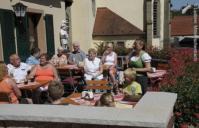 Gasthaus "Zur Krone" Die Gäste des Gasthof Zur Krone sitzen auf der gut gefüllten Steinterassse. Die Terasse ist geschmückt mit roten Blumen. Im Hintegrund ein kleiner Ausschnitt einer Kirche.
