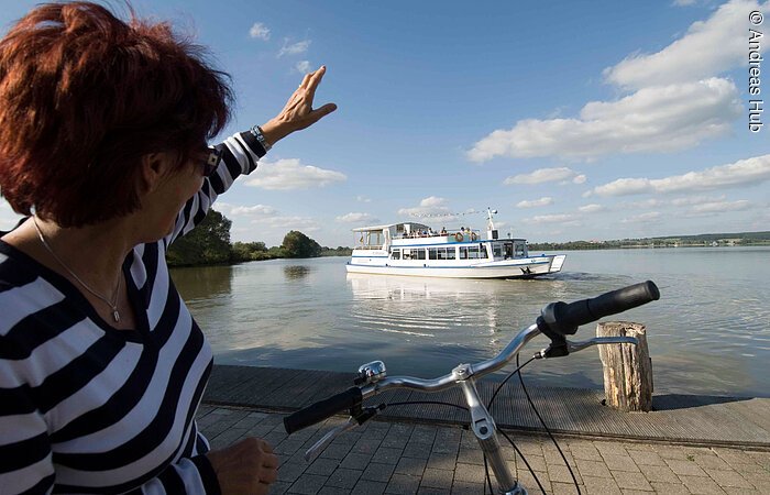 Eine Fahrradfahrerin grüßt die Passagiere der MS Altmühlsee vom Ufer aus Eine Fahrradfahrerin winkt vom Ufer aus der MS Altmühlsee zu.