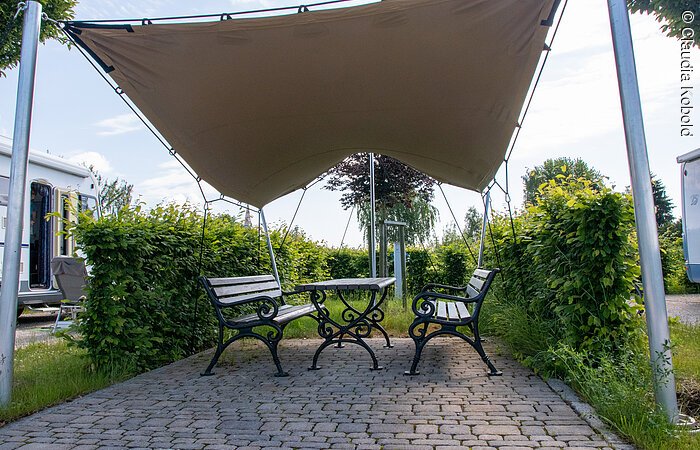 Sitzgelegenheit am Wohnmobilstellplatz in Wolframs-Eschenbach Zwei Bänke und ein Tisch aus Holz stehen unter einem Sonnensegel umgeben von grünen Sträuchern. Im Hintergund erkennt man stehende Wohnmobile.