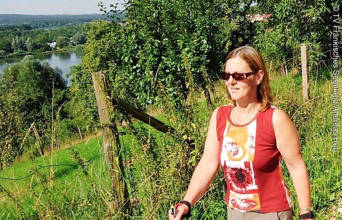 Nordic-Walking Eine Frau in sportlicher Kleidung und Sonnenbrille. Läuft mit Walking-Stöcken in der Hand auf einem Schotterweg entlang einer grünen Wiese. Im Hintergrund sind Bäume und Sträucher. In der Ferne sieht man einen See.