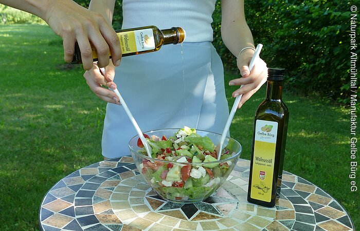 Salat mit Walnussöl Gelbe Bürg Person gießt Walnussöl in eine Schüssel mit Salat auf einem Mosaiktisch im Freien.