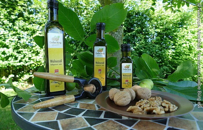 Walnussöl Manufaktur Gelbe Bürg Auf einem runden Mosaiktisch stehen drei Flaschen Walnussöl in verschiedenen Größen mit einem Teller Walnüsse und Walnusskerne. Der Tisch steht vor einem grünen Gebüsch.