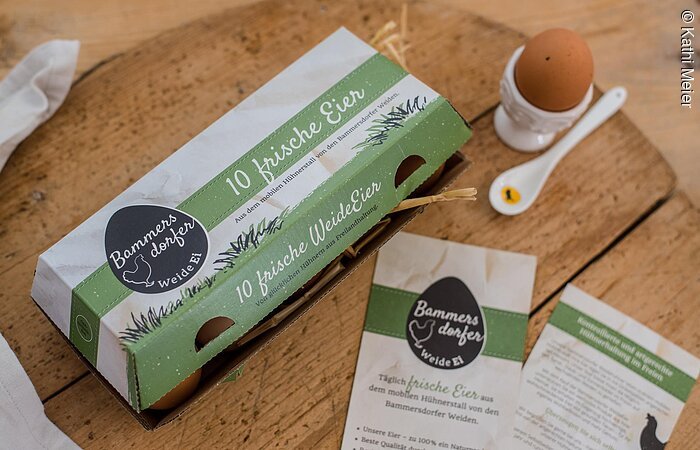 Bammersdorfer Weide Ei Ein geschlossener Eierkarton steht auf einem Holzbrett. Rechts davon ein Eierbecher mit Ei und Löffel. Auf der Eierschachtel steht Bammersdorfer Weide Ei.