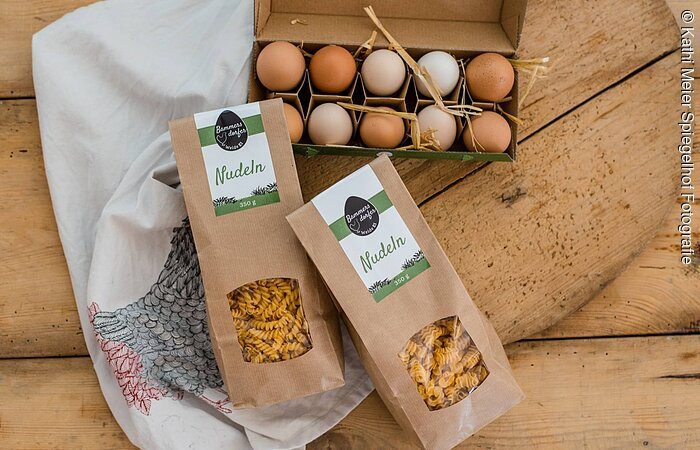 Unsere Eiernudeln Produktpräsentation von Nudeln und Eiern platziert auf Holz. Ein offener, gefüllter Eierkarton. Davor liegen zwei Packungen Nudeln.
