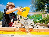 Wasserspielplatz Windsbach Ein Junge auf einem Wasserspielplatz. Er spielt mit einem Wasserspielplatz. Ein gelbes Gefäß aus welchem Wasser tröpfelt.