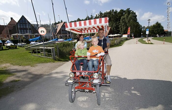Radelspaß am Kleinen Brombachsee©TV Fränkisches Seenland/TV Franken/A. Hub Eine Ehepaar in einer Rikscha radelt mit ihren zwei Kindern auf einem Fahrradweg. Sie fahren vorbei an einem großen Haus mit Trockenliegeplätzen für Segelboote.