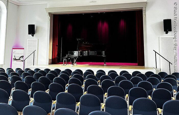 Der Wildbadsaal in Weißenburg (Bay) Leerer Konzertsaal mit Reihen blauer Stühle und einem Flügel auf der Bühne unter einer Deckenleuchte.